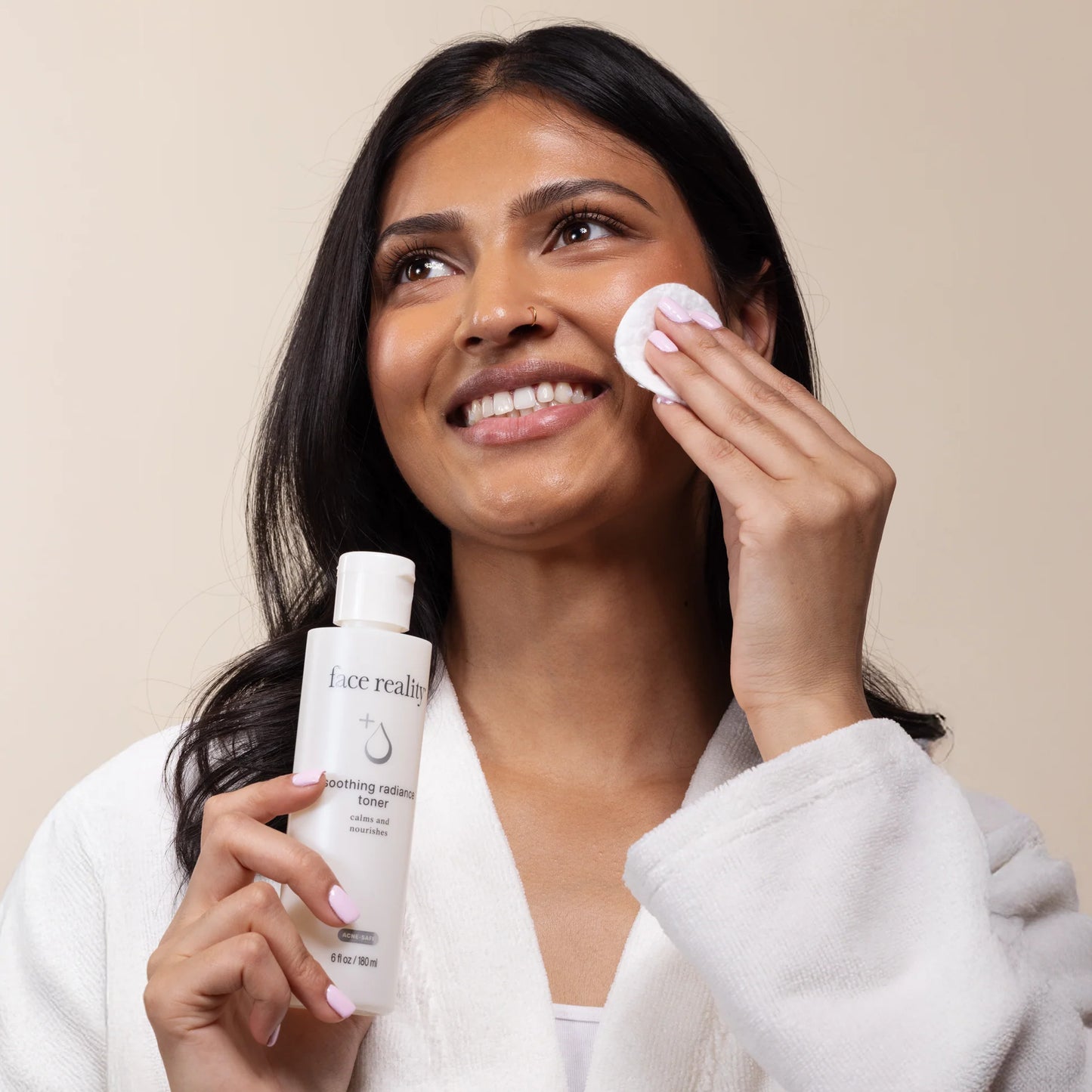 Woman applying skincare product with a bottle of 'Face Reality' soothing radiance toner on a beige background