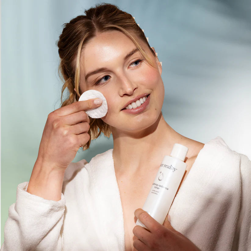 Woman applying skincare product with a cotton pad and holding a face reality bottle against a light background